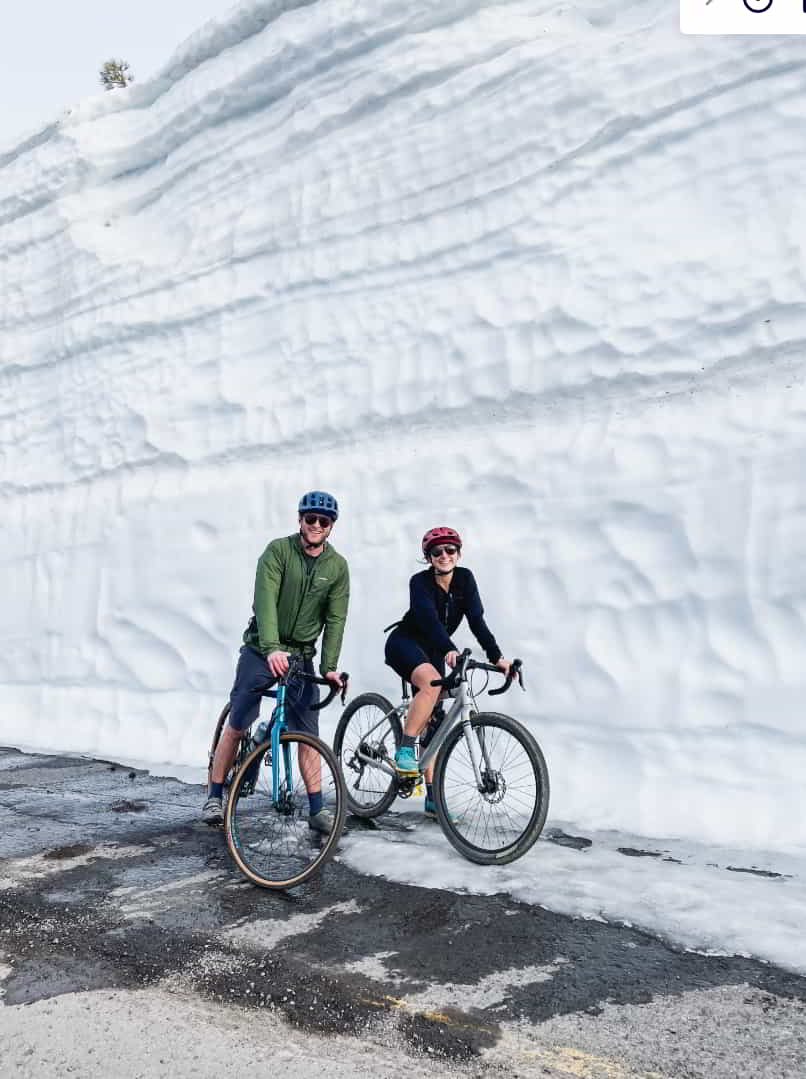 2 people posing on their bikes in front of a wall of snow next to the road